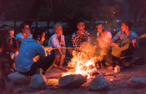 young millennial couples listening to guitar around campfire at cabins and lake