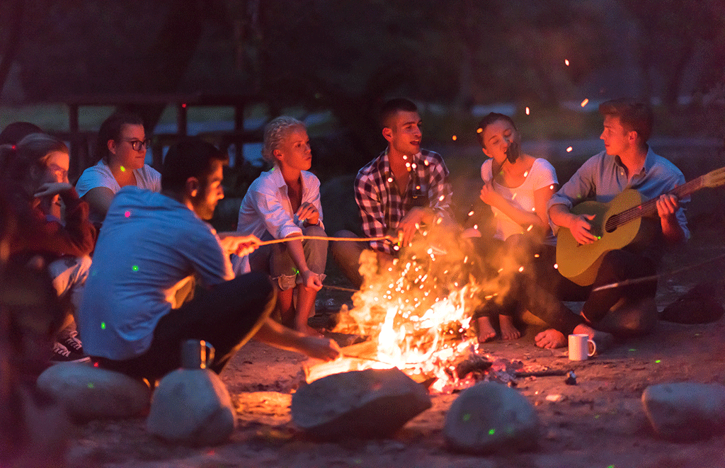 young millennial couples listening to guitar around campfire at cabins and lake